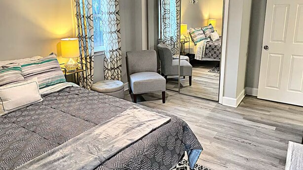 A bedroom with a large bed, a chair, a nightstand, and a mirror. - Charming Coastal Cottage Near Beach & Downtown (Wilmington)