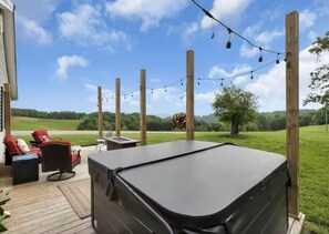 A covered outdoor deck with a hot tub, red chairs, and string lights against a backdrop of a grassy field and trees. - Ridgeview Cottage - 6 miles to the Rock House! (Laurelville)