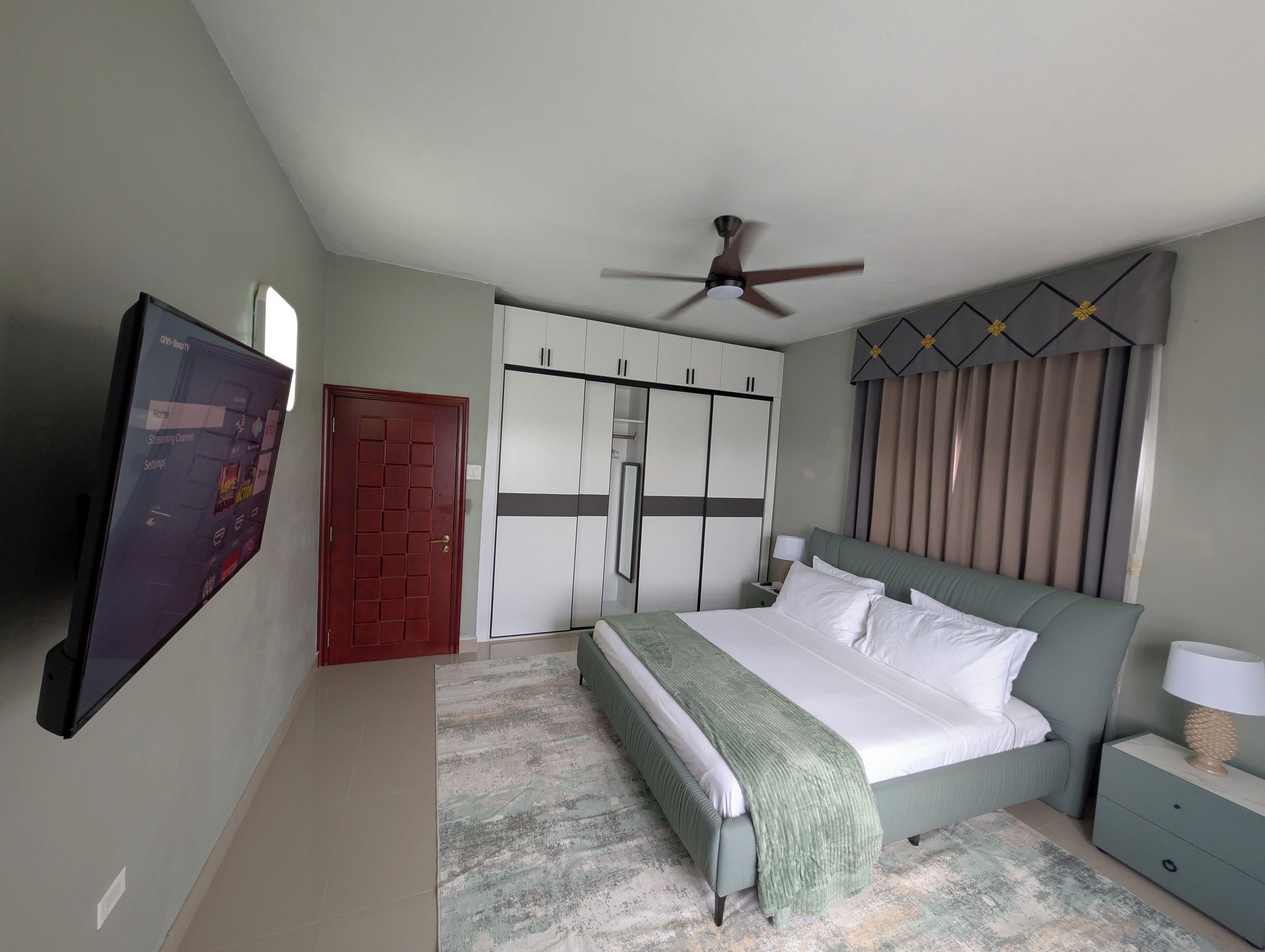 Ein Schlafzimmer mit Bett, Deckenventilator, Kleiderschrank, Fernseher und einer Tür.