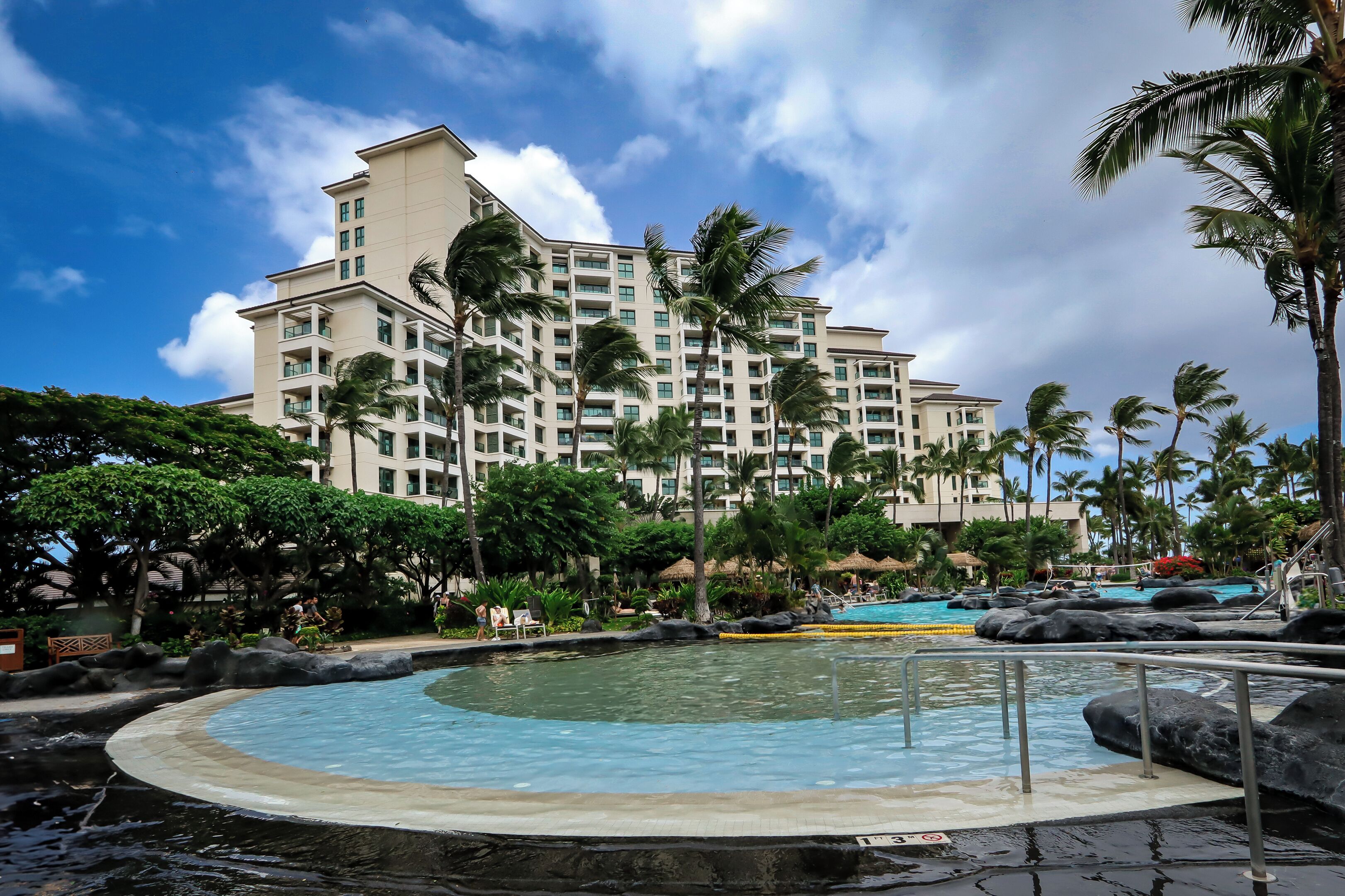 A hotel with a large pool, palm trees, and a clear sky.