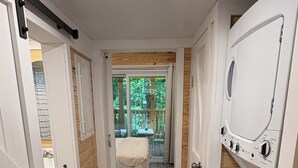 A compact, modern room with a wooden floor, a bed with a view of the outdoors, and a built-in washing machine. - New cottage with Hot Tub, Tiki Bar, Sauna, and Plunge Tub. Million Dollar Views (Brevard)