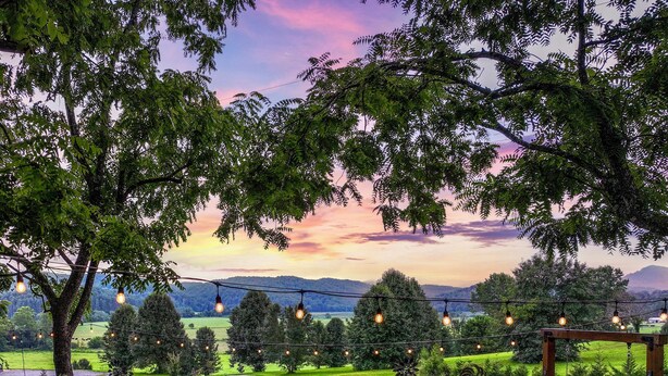 An outdoor seating area with Adirondack chairs and a swing, overlooking a scenic landscape with trees and a sunset. - New cottage with Hot Tub, Tiki Bar, Sauna, and Plunge Tub. Million Dollar Views (Brevard)