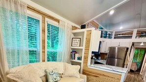 A modern living room with a sofa, a coffee table, and a kitchen area with a refrigerator and wooden cabinets. - New cottage with Hot Tub, Tiki Bar, Sauna, and Plunge Tub. Million Dollar Views (Brevard)