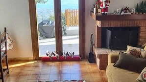 En stue med en sofa, et sofabord dækket af en rød dug, en pejs og udsigt gennem en glasdør.