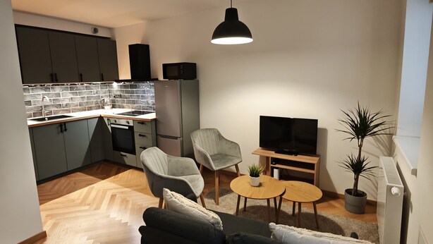 A modern living room with a sofa, a coffee table, and a TV. There is a kitchen area with cabinets and appliances in the background.