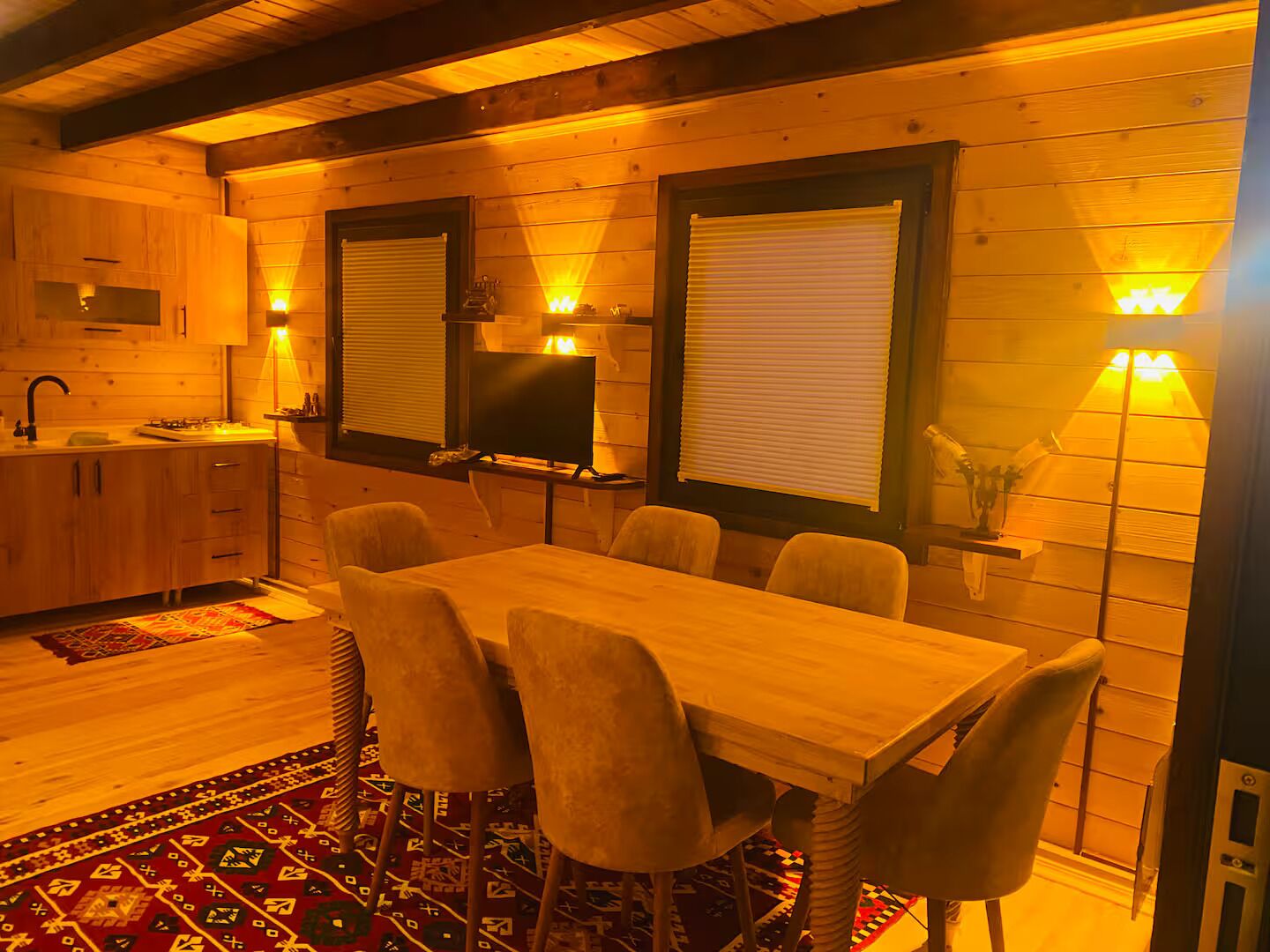 A cozy wooden cabin interior with a dining area, kitchen, and two windows with blinds.