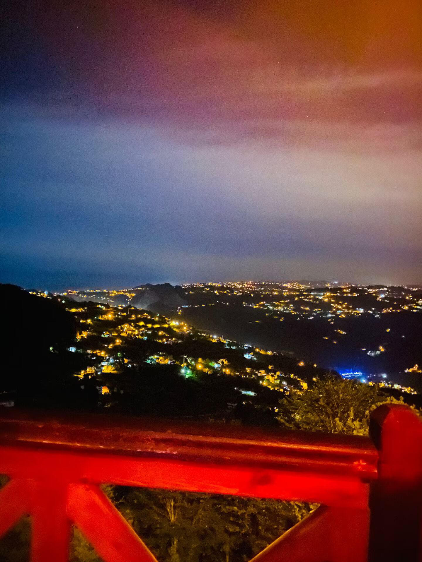 A nighttime view of a city from a high vantage point, featuring a red railing in the foreground.