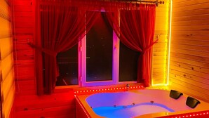 A room with a wooden floor, a large window with red curtains, a jacuzzi with LED lights, and a small wooden chest. - Of Kartepe Bungalov & İkiz Villalar (Of)