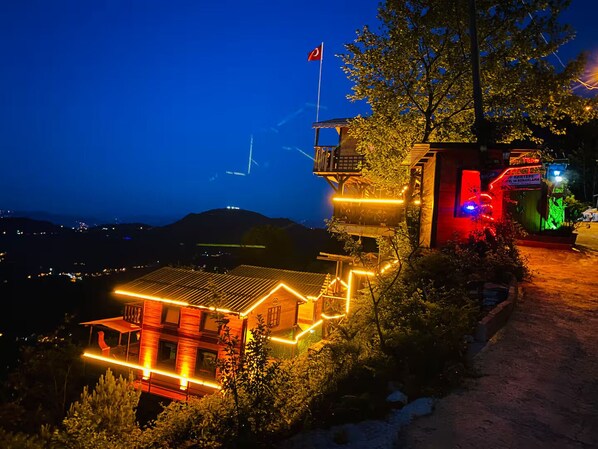A building with a flag, illuminated by warm lights, surrounded by trees and a view of the landscape. - Of Kartepe Bungalov & İkiz Villalar (Of)