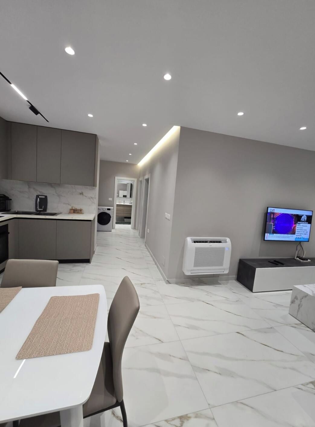A modern kitchen with a dining area, a TV, and a hallway leading to a bathroom.