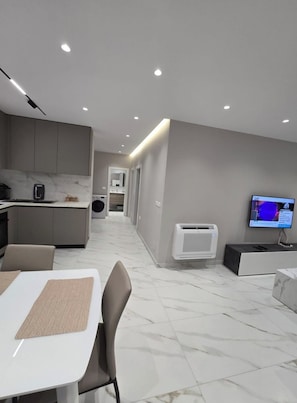 A modern kitchen with a dining area, a TV, and a hallway leading to a bathroom.
