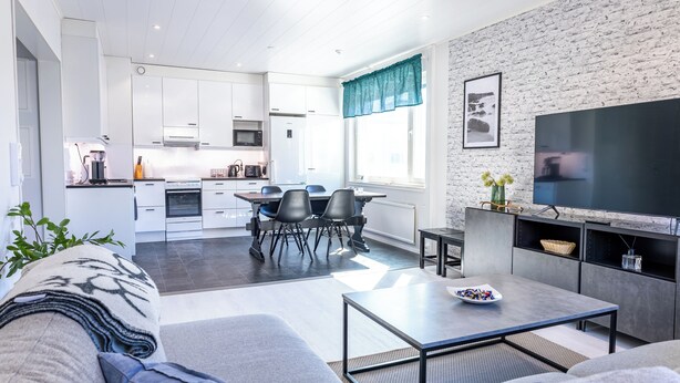 A modern living room with a sofa, a coffee table, and a TV. There is a dining area with chairs and a table. The kitchen is visible in the background with white cabinets and a microwave.