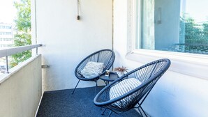 A balcony with two wicker chairs and a small table, a white wall, and a window overlooking a building.