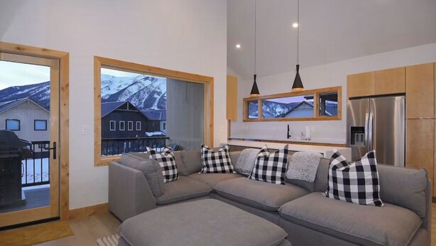 Un salon moderne avec un canapé d’angle gris, des meubles en bois et une grande fenêtre donnant sur les montagnes.