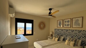 A bedroom with two beds, a ceiling fan, a window with a view of buildings, and framed artwork on the wall. - Tortuguero vacation apartment: air conditioning and sea views, ideal for couples (Morro Jable)