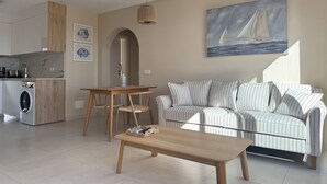 A modern living room with a striped sofa, a wooden coffee table, and a dining table with chairs. There is a kitchen area with a washing machine and a sink. A painting of sailboats hangs on the wall. - Tortuguero vacation apartment: air conditioning and sea views, ideal for couples (Morro Jable)