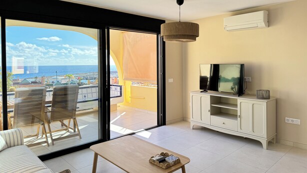 A modern living room with a large sliding glass door leading to a balcony, a flat-screen TV, a white cabinet, and a wooden table. - Tortuguero vacation apartment: air conditioning and sea views, ideal for couples (Morro Jable)