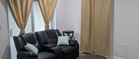 Un salón con un sofá de cuero negro, una mesa de centro, cortinas color beige y un reloj de pared.