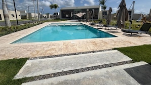 A rectangular swimming pool with a tiled deck, surrounded by lounge chairs and umbrellas, with a modern building in the background. - The Windown Suite Near Downtown Punta Cana (Higuey)