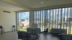 A spacious indoor area with large windows, wicker furniture, and a view of a swimming pool and buildings. - The Windown Suite Near Downtown Punta Cana (Higuey)