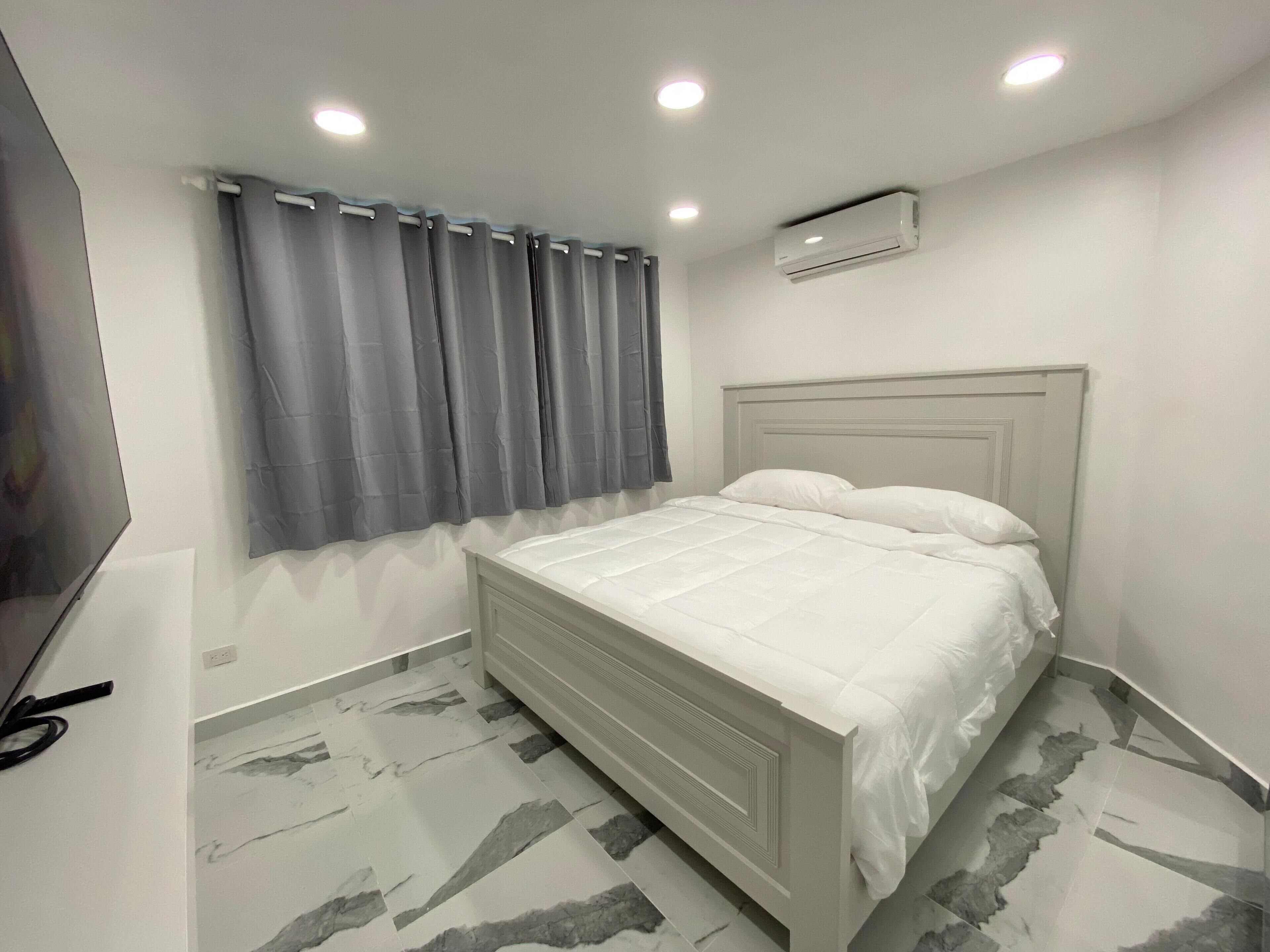 A bedroom with a bed, grey curtains, an air conditioning unit, and a TV.