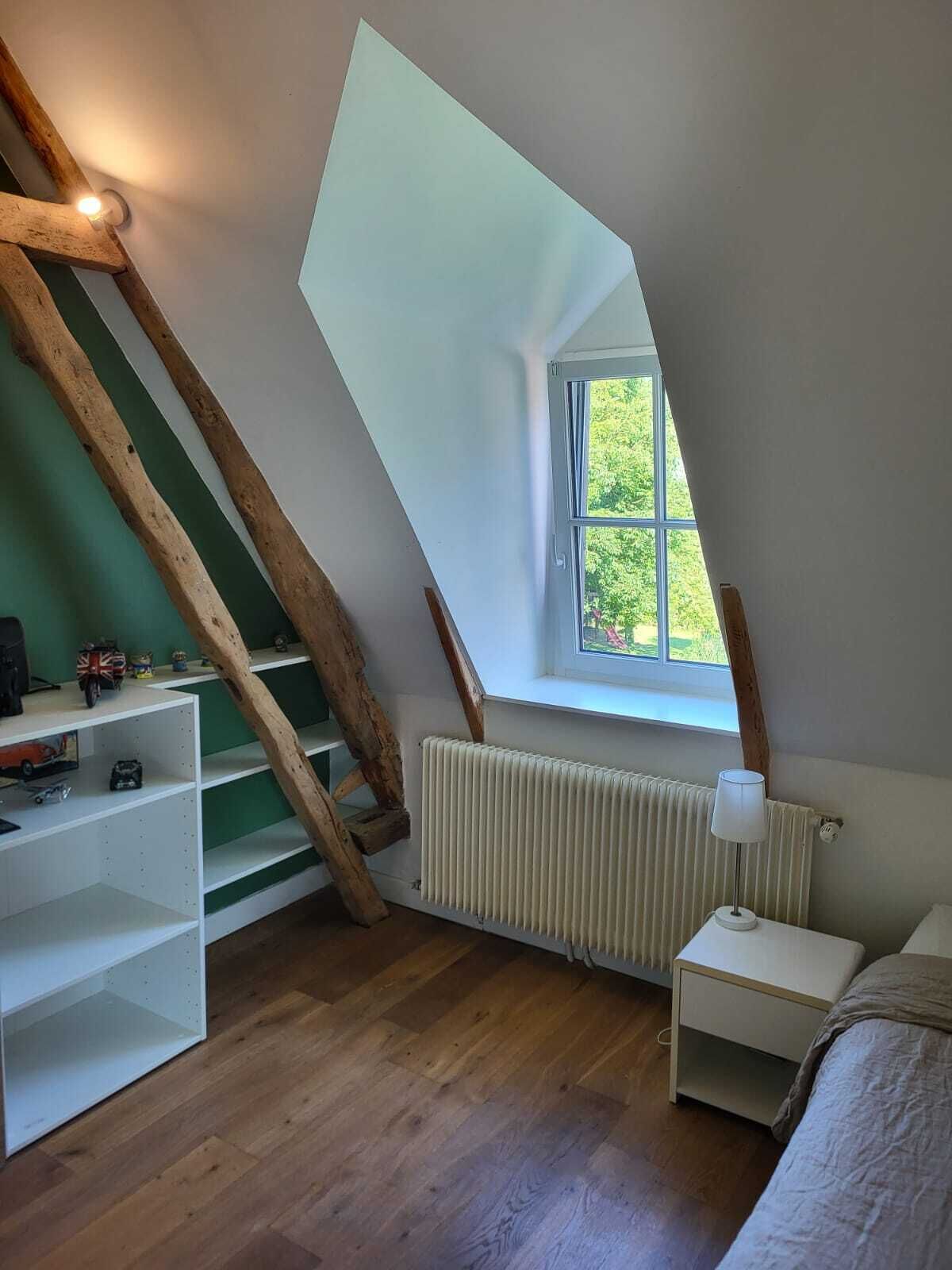 A room with a sloped ceiling, a window with a view of greenery, a radiator, a white shelf unit, and a bedside table with a lamp.