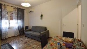 A living room with a sofa, a dining table with a floral tablecloth, a small plant on the wall, and a window with curtains. - B&b on the course, Reggio Calabria (Reggio Calabria)