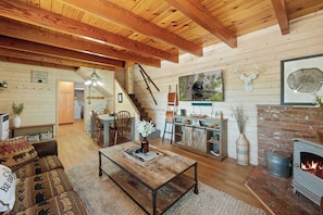 A rustic cabin interior with a wooden ceiling, a fireplace, a dining area, and a living room with a sofa and coffee table.