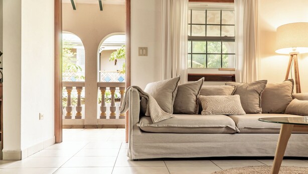 En stue med en beige sofa, et glasbord og en lampe. Udsigt til en balkon med planter og gelænder.
