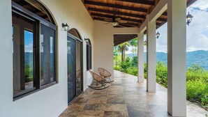 A covered outdoor area with a tiled floor, two rocking chairs, and a view of a lush landscape. - Getaway Charming 2-bedroom house secluded with Ocean Breeze (San José Province)