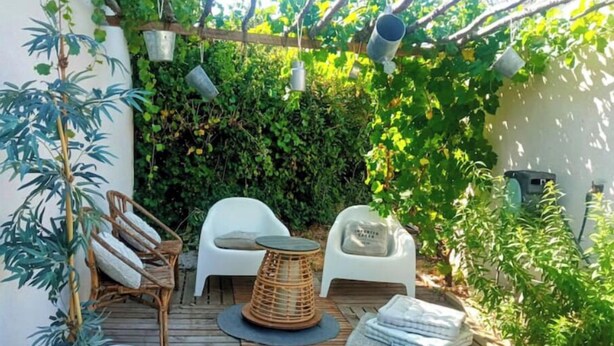 Une terrasse en bois, meublée de meubles en osier, de chaises en plastique blanc et d’un pot en métal.