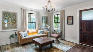 A living room with a sofa, coffee table, and a chandelier. There are windows with curtains and a door. - Downtown Mobile: Pet Friendly + 5m Walk (Mobile)