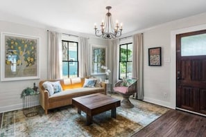 A living room with a sofa, coffee table, and a chandelier. There are windows with curtains and a door.