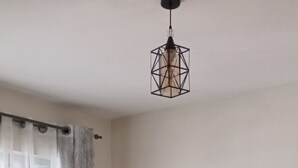 Ein Schlafzimmer mit Bett, einem Fenster mit Vorhängen, einer Pendelleuchte und einer Wandsteckdose.