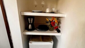 A compact kitchenette with a white refrigerator, a coffee maker, and a small shelf with wine glasses and a vase of flowers. - Casa Brisa Noronha (Fernando de Noronha)