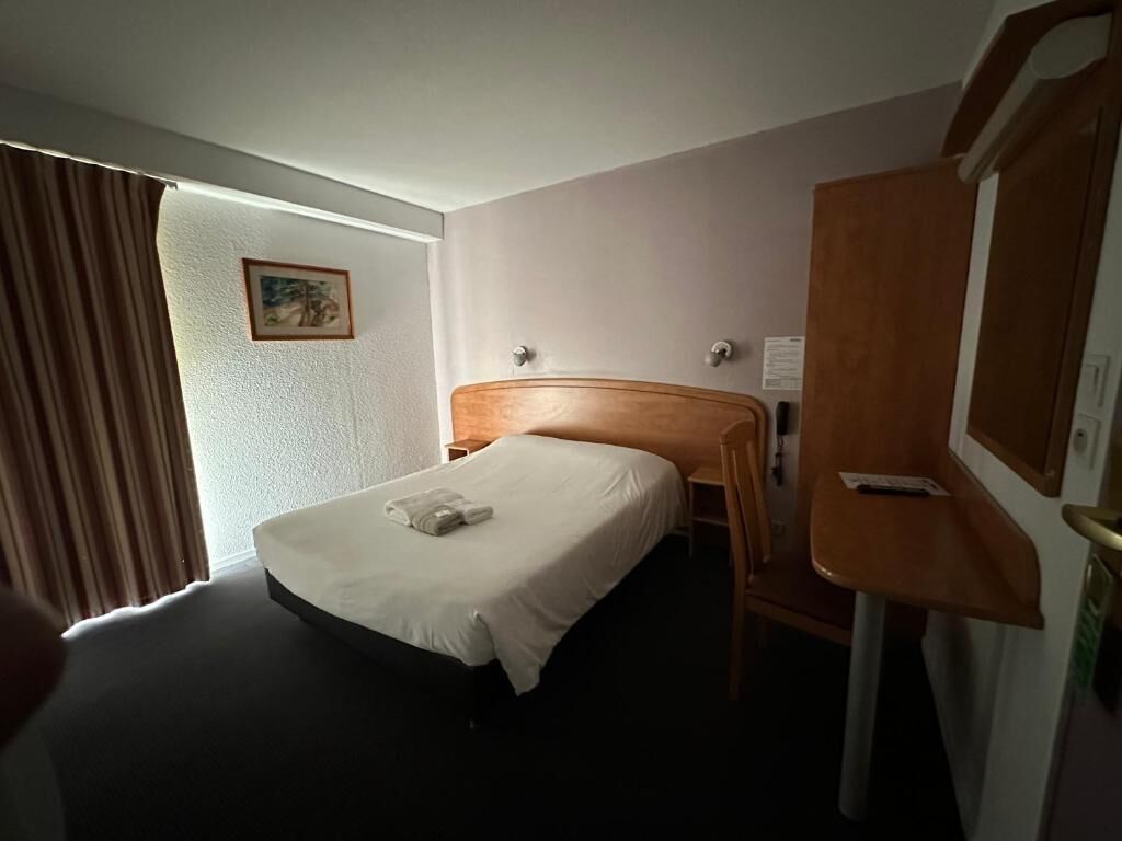Habitación de hotel con cama, cabecero de madera, escritorio con silla, espejo y ventana con cortinas.