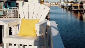 A waterfront property with a dock, a white chair with a yellow cushion, and a row of buildings along the water. - Upscale Waterfront Boat Lift~Kayaks~Pets~Sleeps12 (Galveston)