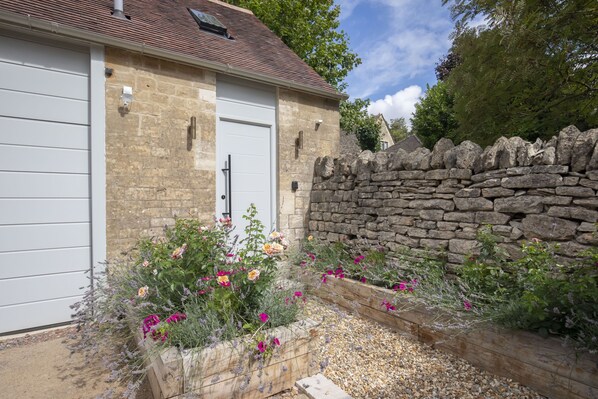 A stone building with a white garage door, surrounded by a stone wall and a garden with various flowers.