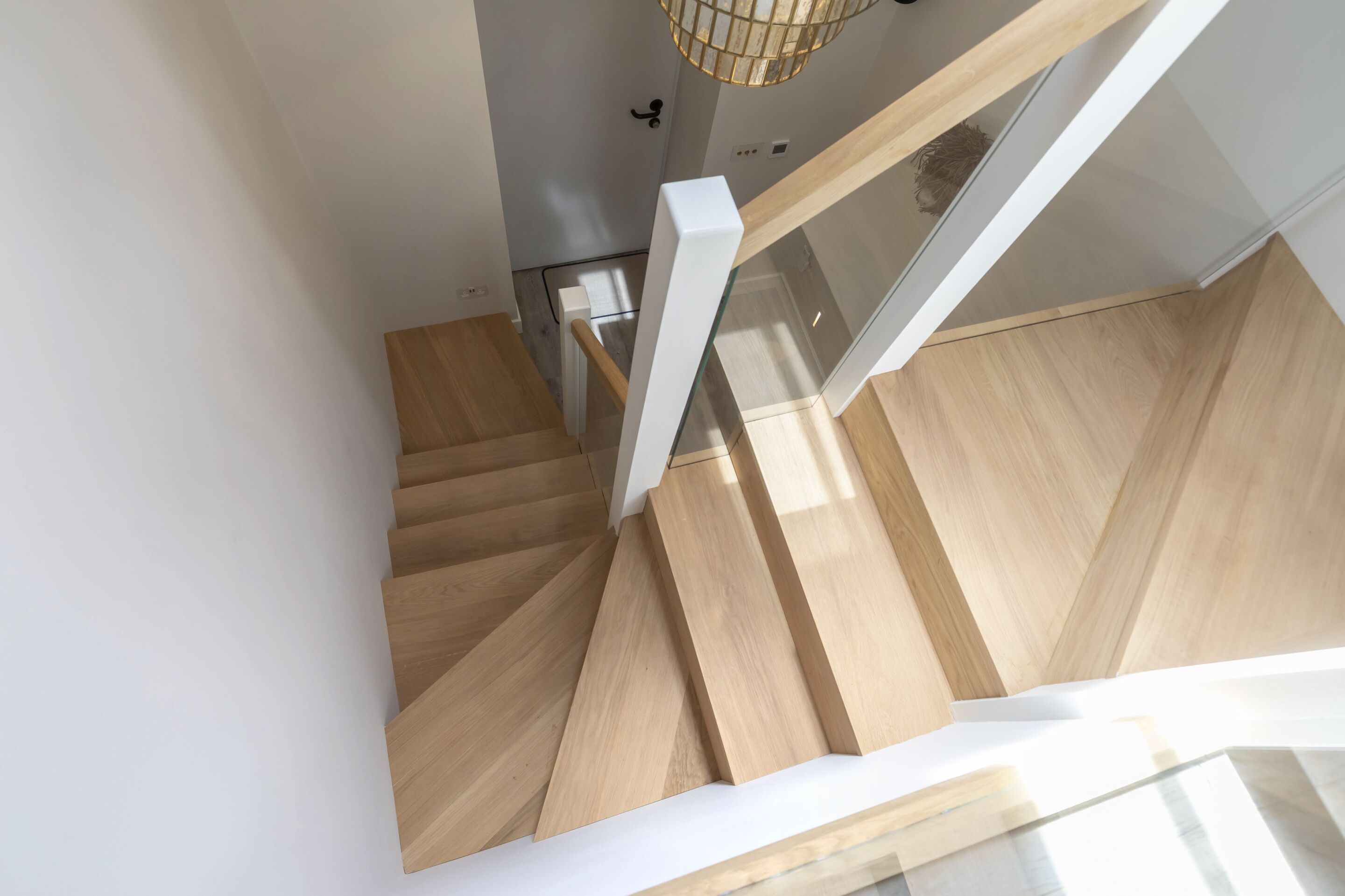 A wooden staircase with white handrails and a glass railing, leading to a room with a white door and a hanging light fixture.