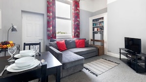 A modern living room with a grey sofa, red cushions, a dining table set with plates and glasses, a bookshelf, and a TV.