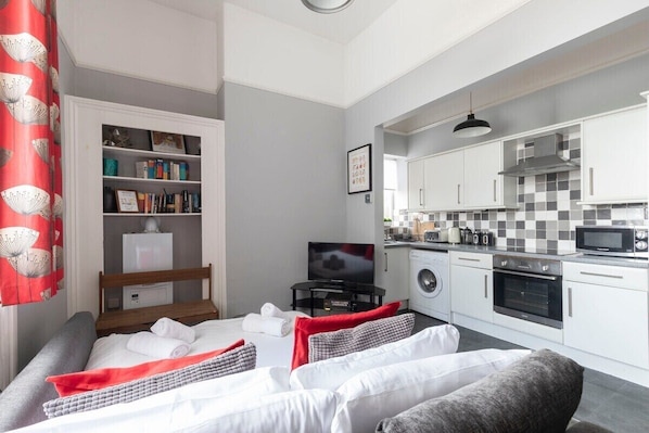 A modern hotel room with a bed, a small kitchenette, a bookshelf, and a television.