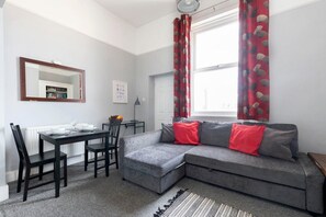 Un salon avec un canapé d’angle gris, une table à manger avec des chaises, un miroir et une fenêtre avec des rideaux rouges.