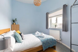 Ein Schlafzimmer mit Bett, Nachttisch mit Lampe, Fenster mit Vorhängen und Heizkörper.