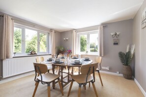 A dining room with a round table set for four, surrounded by chairs. There are windows on two sides, a radiator, and a wall-mounted light fixture. - A Quaint, Relaxing 3-Bed Cottage in Eastington (Gloucestershire)