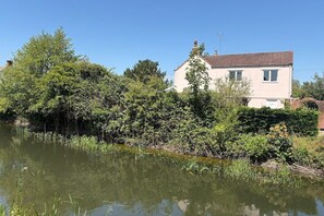 Une maison au bord d’un plan d’eau paisible, entourée d’une végétation luxuriante et sous un ciel dégagé.