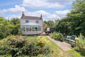 A house with a conservatory, surrounded by a garden with various plants and a bench. - A Quaint, Relaxing 3-Bed Cottage in Eastington (Gloucestershire)