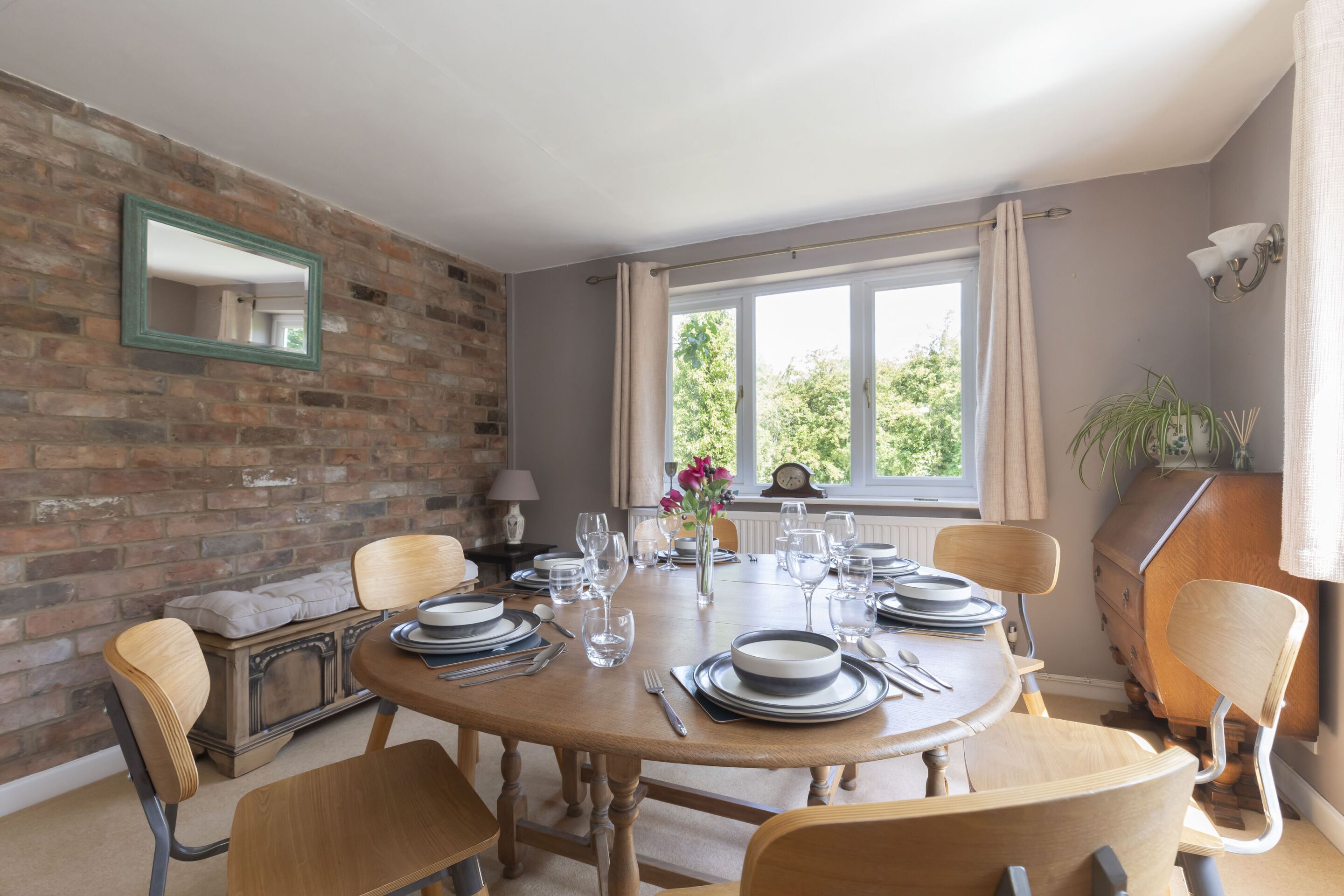 A dining room with a brick wall, a round wooden table set for six, and a window with curtains.