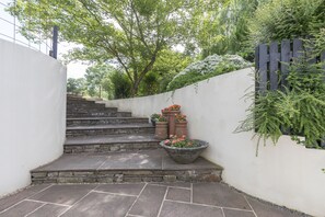 Un escalier en pierre menant à un mur blanc, surmonté d’une clôture en bois et agrémenté de verdure.