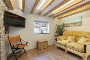 Une pièce avec un plancher en bois, un canapé agrémenté de coussins à motifs, une chaise en bois, un téléviseur fixé au mur, une fenêtre donnant sur un paysage verdoyant et un tableau accroché au mur.