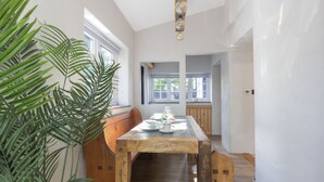 Un coin repas avec une table en bois dressée pour quatre personnes, entourée de grandes fenêtres et agrémentée d’une plante en pot.
